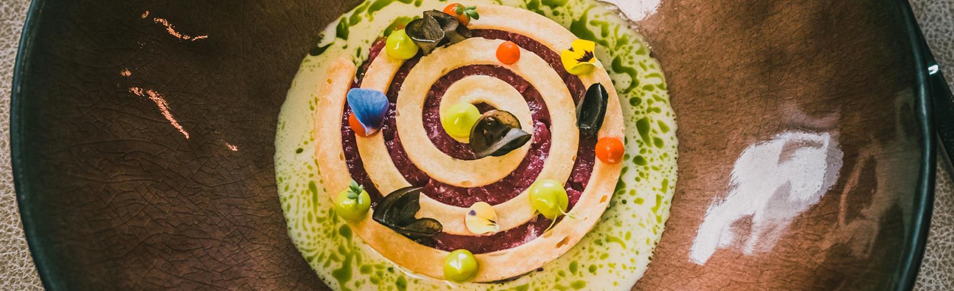 Kunstvoll angerichtete Vorspeise mit Spirale, Tropfen und Blüten auf braunem Teller.
