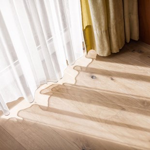Sonnenlicht fällt durch weiße Gardinen auf einen hellen Holzboden neben einem Vorhang.
