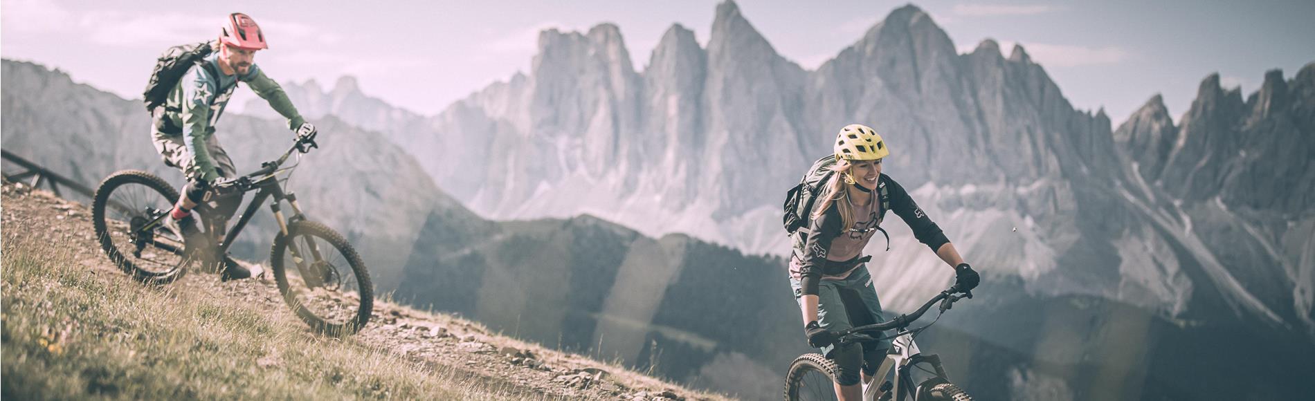 Due ciclisti in MTB su un sentiero con le Odle delle Dolomiti sullo sfondo.