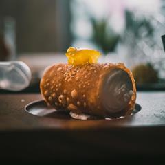 Close-up of a crispy cannolo filled with cream and topped with a drop of yellow jam on a wooden tray.