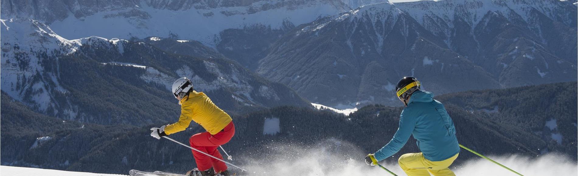 Zwei Skifahrer auf frisch präparierter Piste mit Blick auf verschneite Berge im Hintergrund.