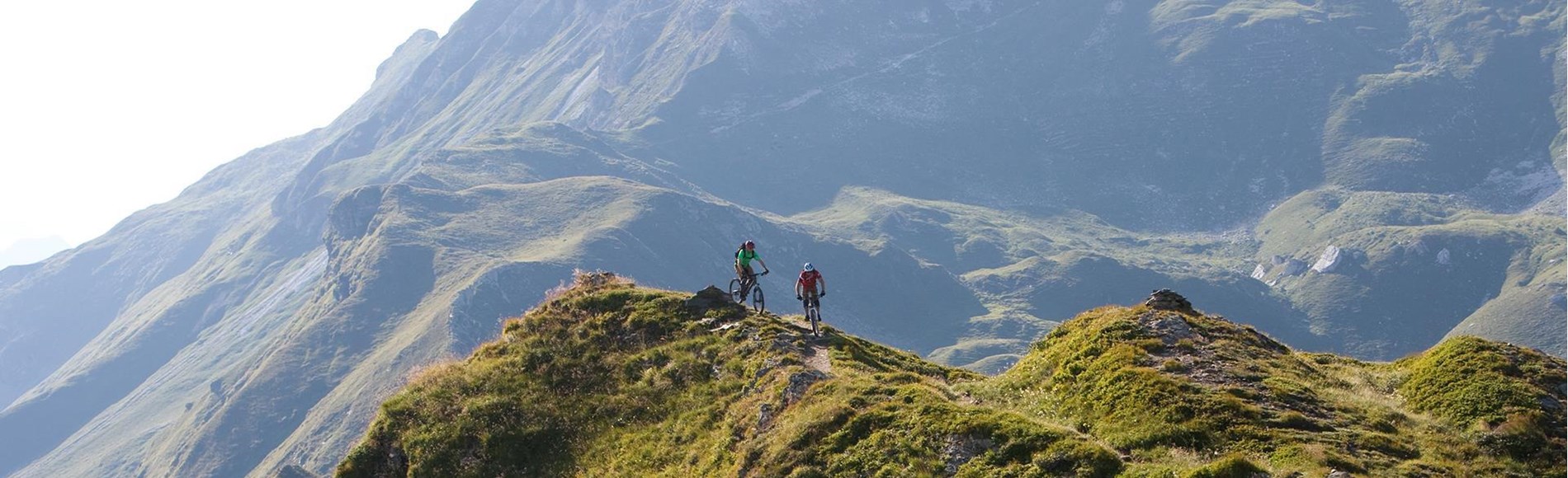 Zwei Mountainbiker fahren auf einem schmalen Gratweg inmitten einer beeindruckenden Berglandschaft.