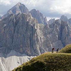 Two mountain bikers on a high trail with massive Dolomite cliffs towering in the background.