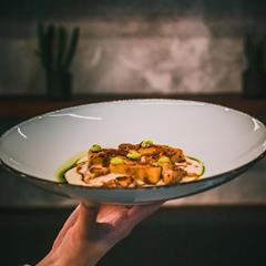 A hand holds a plate with roasted pumpkin, creamy sauce and green drops against dark background.
