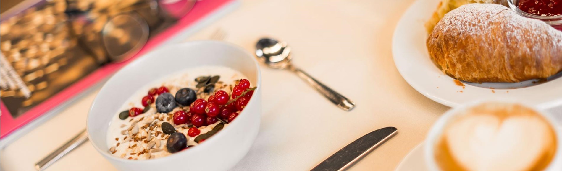 Colazione con yogurt, cornetto, caffè e panini su una tavola apparecchiata.